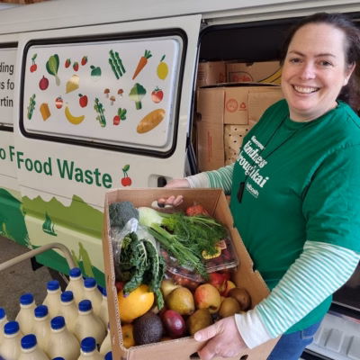 Woman holding box of produce stands in front of a Kaibosh food rescue van next to crates of milk bottles.