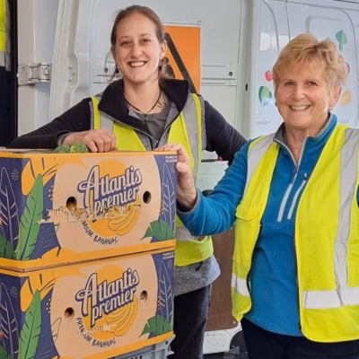 Two women stand beside a food rescuevan unloading a trolley of food in banana boxes.