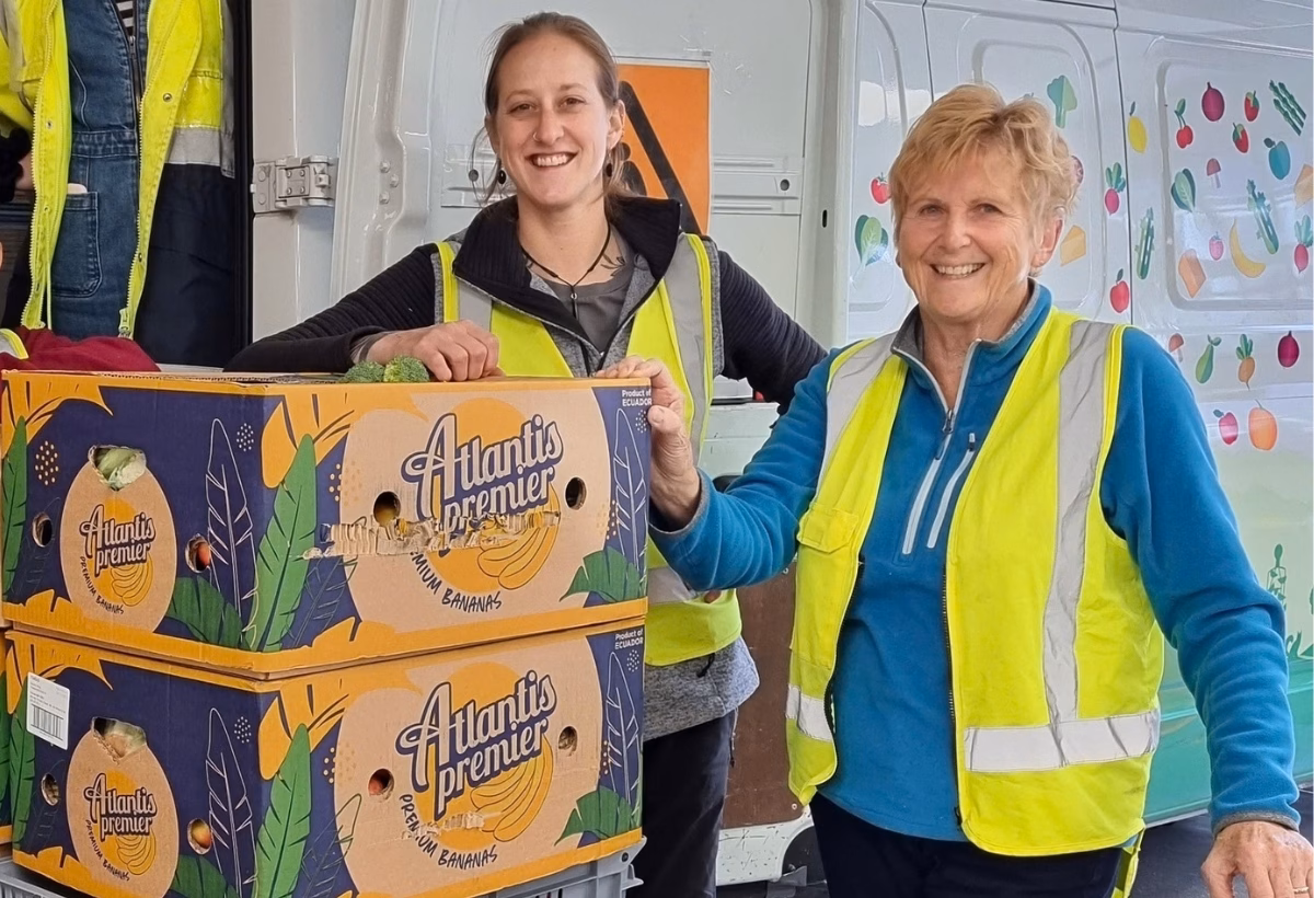 Two women stand beside a food rescuevan unloading a trolley of food in banana boxes.